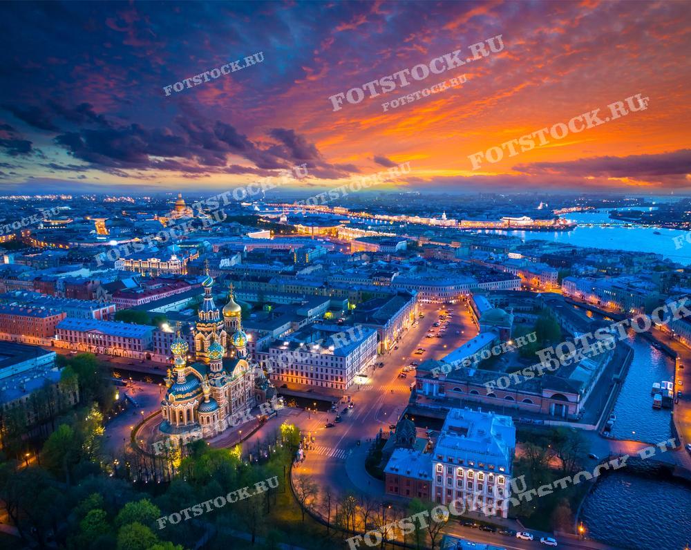 На фотографии — вид на Санкт-Петербург в момент заката или рассвета. Центром композиции является Храм Спаса на Крови с его яркими цветными куполами, которые выделяются на фоне городских огней и вечернего неба, окрашенного в огненные тона. Небо с заходящим солнцем создает захватывающее зрелище с облаками, переливающимися от оранжевого до фиолетового. Разноцветное освещение подчеркивает историческую архитектуру и добавляет фотографии драматизма. Изображение идеально подойдет для использования в печатных и онлайн публикациях, посвященных культуре и истории Санкт-Петербурга, для туристических проспектов и как украшение интерьеров.